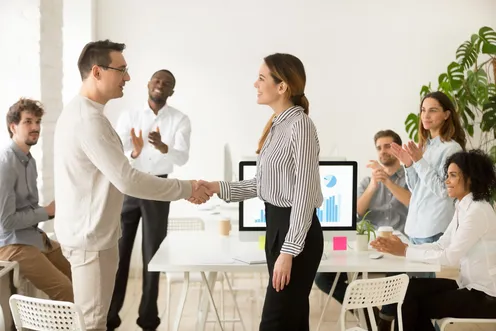 A new colleague greets a teammate with a handshake as coworkers applaud in an office.